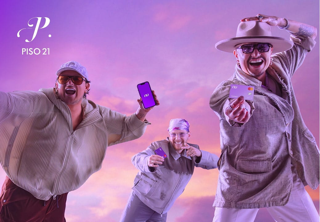 Tres integrantes del grupo musical Piso 21 posan para la cámara, con fondo de atardecer morado. Uno de ellos, en el centro, sostiene un teléfono morado con la aplicación de Nubank abierta, mientras los otros dos, en los costados, sostienen tarjetas moradas de Nubank.