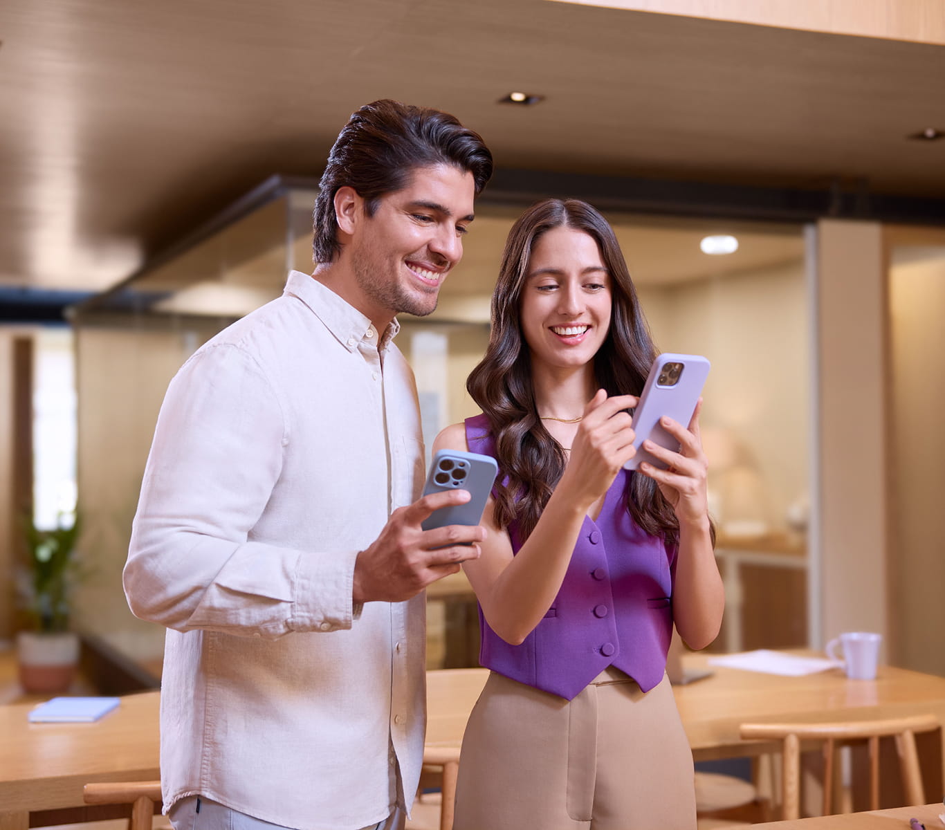Un hombre con camisa blanca y un mujer con chaleco morado, indicando algo en su celular, ambos sonriendo mientras observan el celular de la mujer. Un hombre con camisa blanca y un mujer con chaleco morado, indicando algo en su celular, ambos sonriendo mientras observan el celular de la mujer.